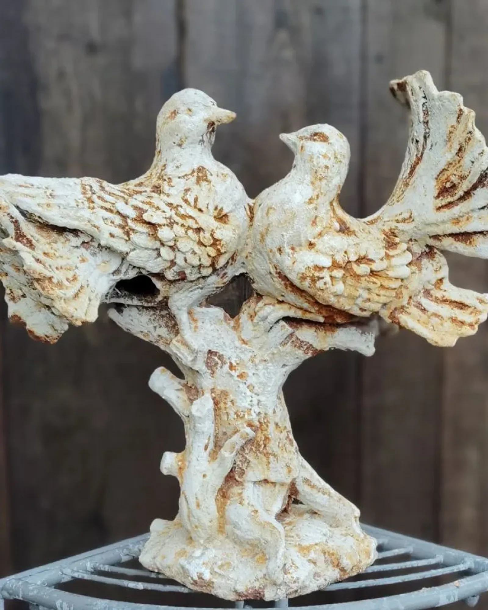 Two cast iron doves perched side by side on a branch, in a distressed cream finish with rust showing through, on a grey metal stand.
