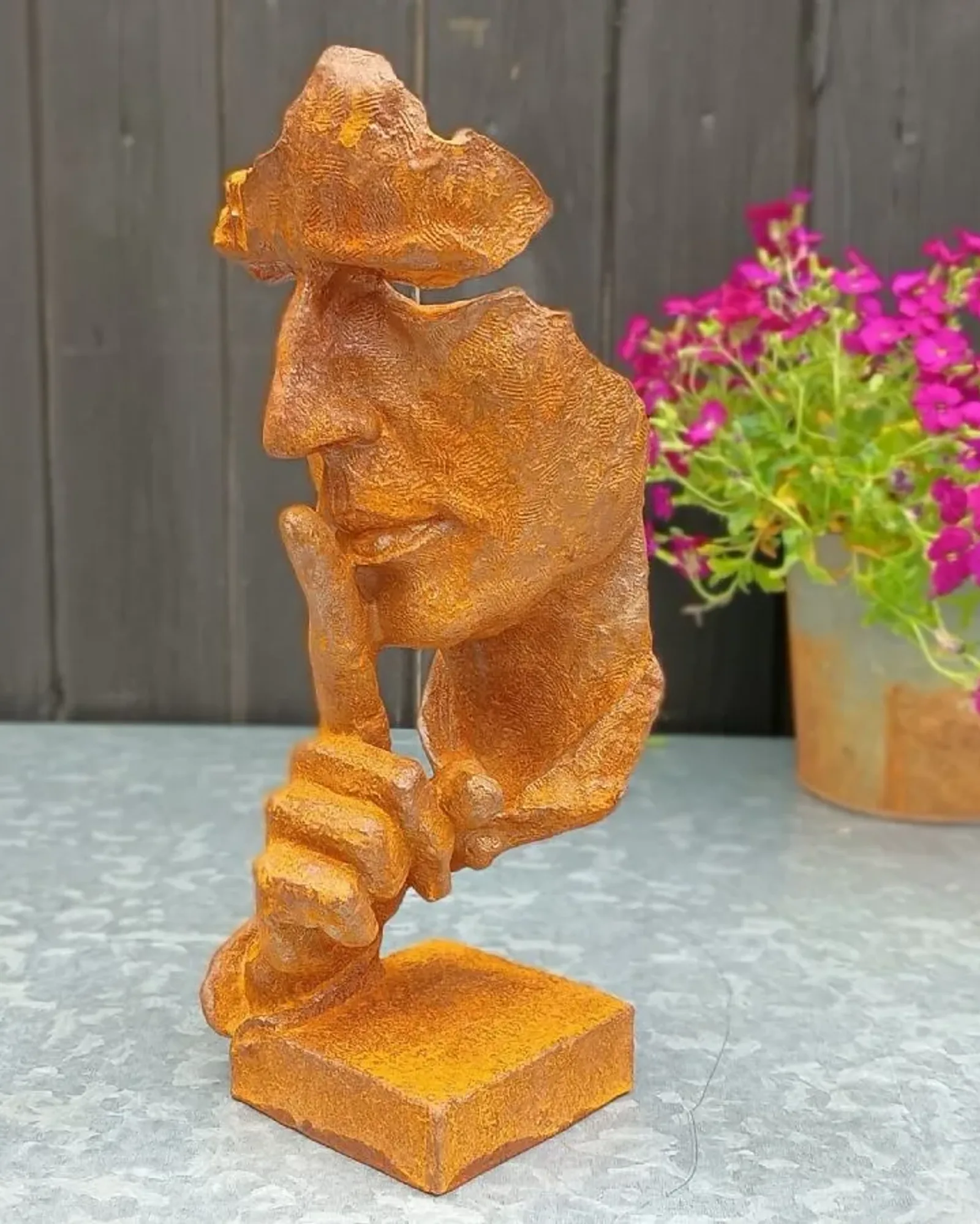 A rusted cast iron sculpture of a stylised face with one finger held to its lips, standing on a square base with a flowering pot behind.