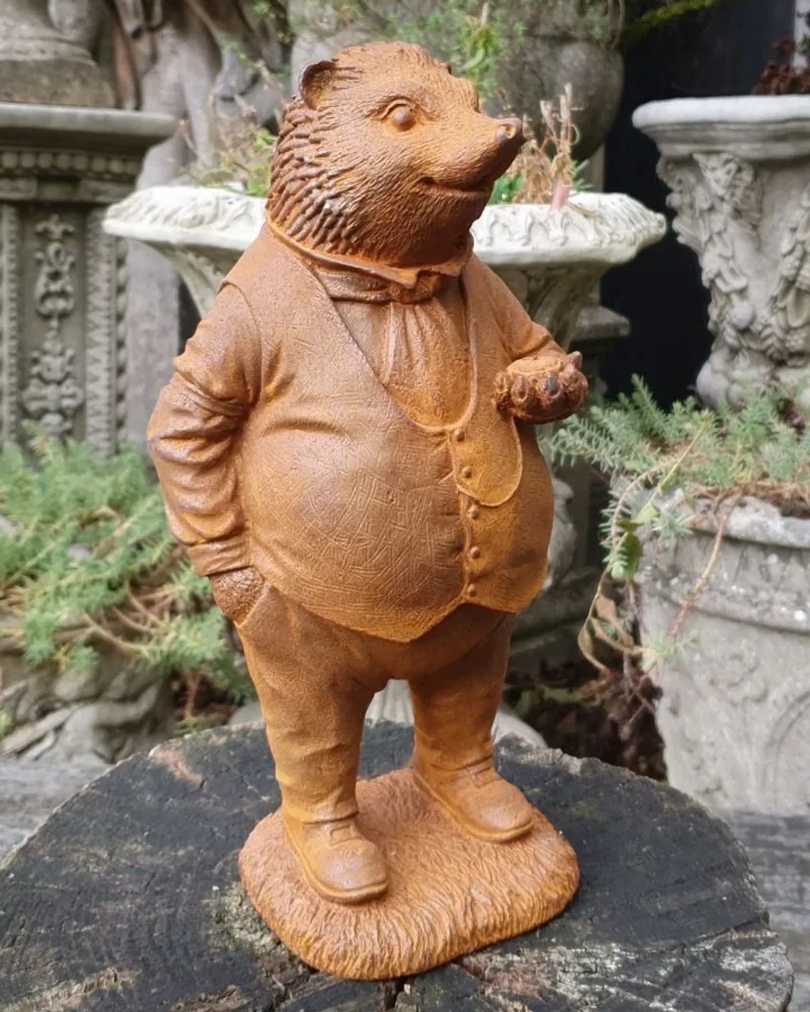 A rusted cast iron hedgehog standing upright in a waistcoat and cravat, holding an acorn, on a grassy base on a log.
