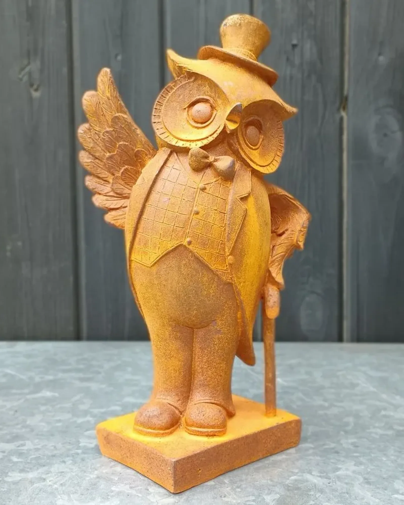 A rusted cast iron owl in a top hat, check waistcoat and bow tie, holding a cane, on a rectangular base against a grey timber wall.