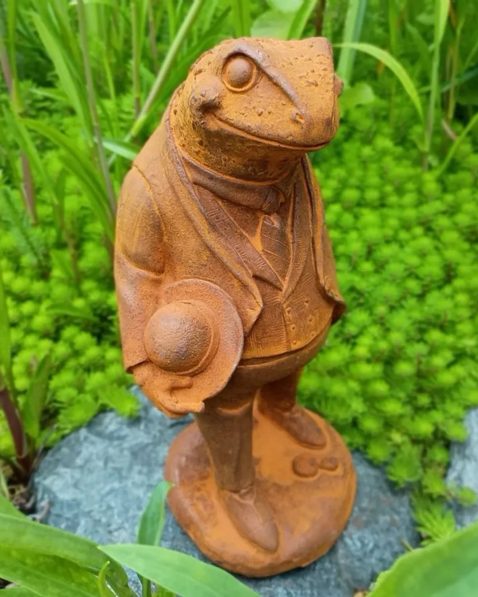 A rusted cast iron toad standing upright in a jacket and waistcoat, holding a bowler hat, on a round base among greenery.