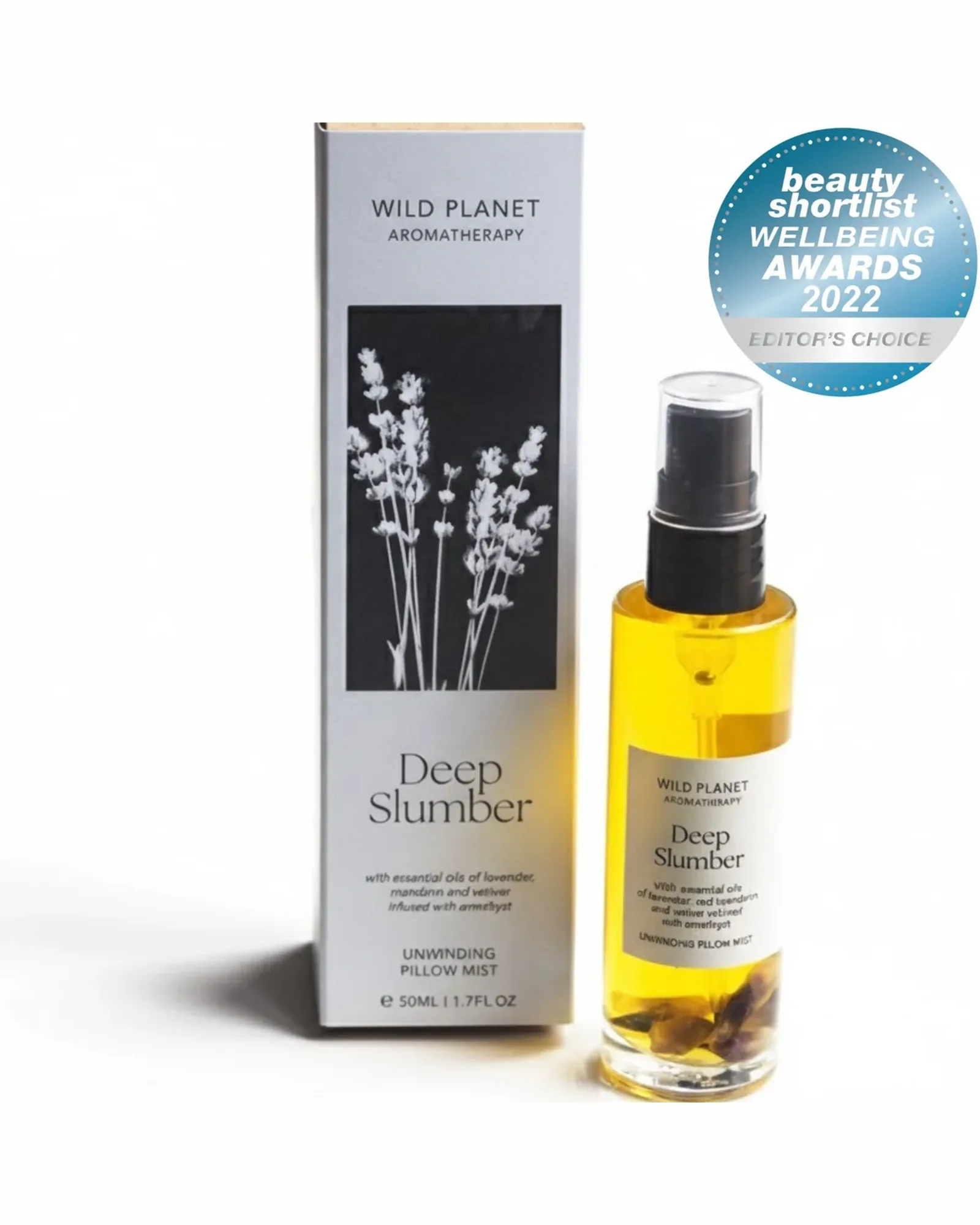 A slim clear glass bottle of Deep Slumber pillow mist with a black spray pump and small amethyst chips at the bottom, beside its card box printed with lavender.