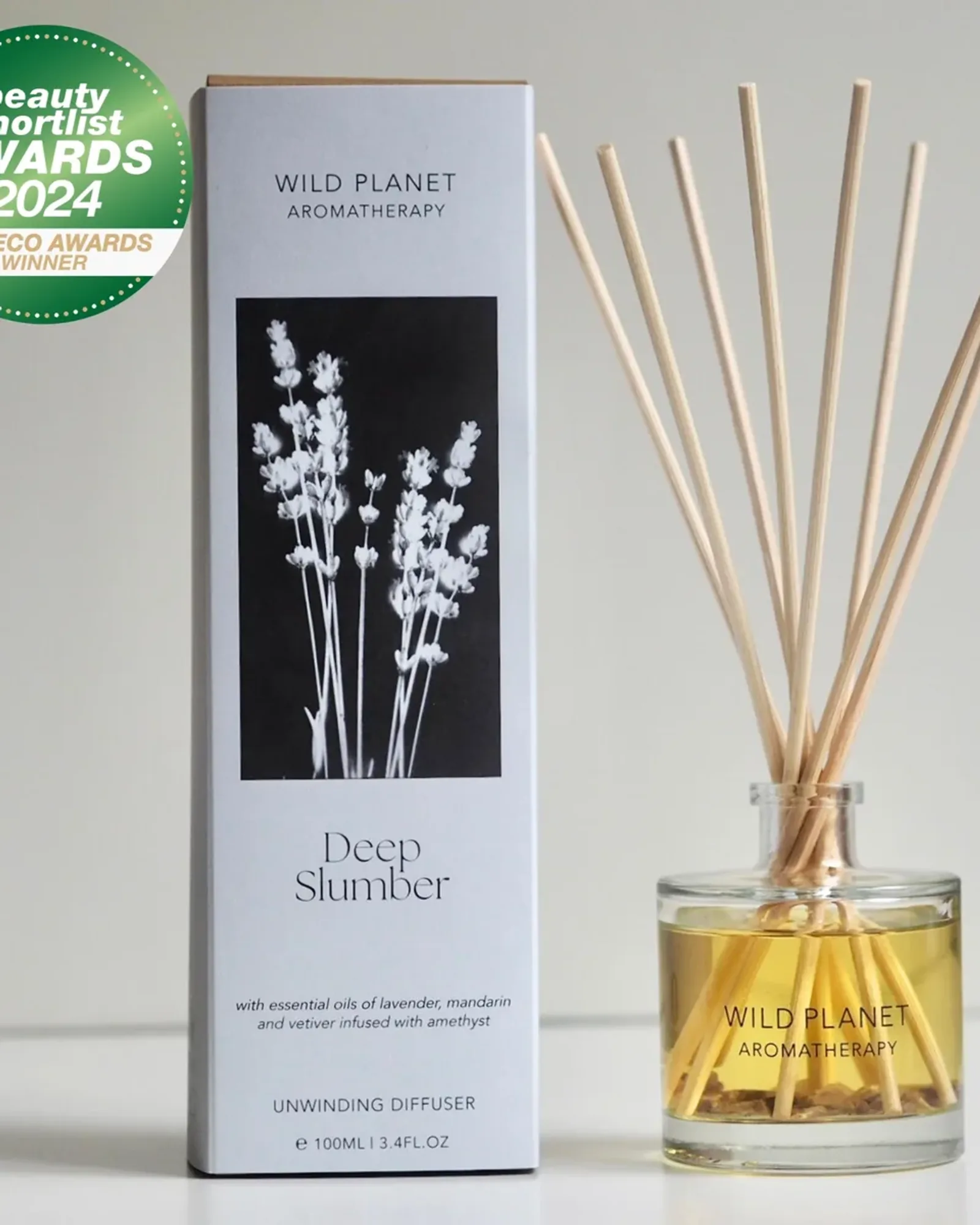 A clear glass bottle of Deep Slumber reed diffuser oil with a fan of natural reeds standing in it, beside its card box printed with the scent's name.