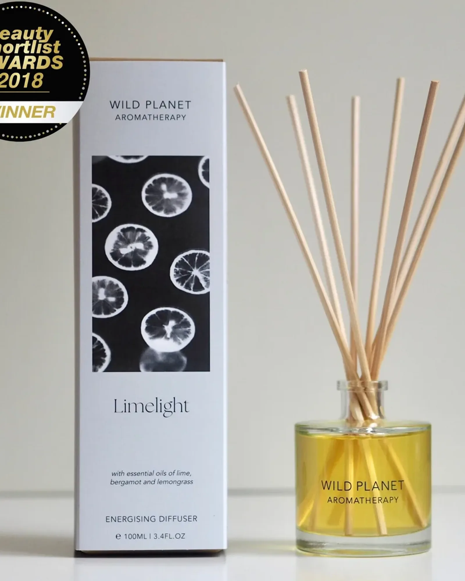 A clear glass bottle of Limelight reed diffuser oil with a fan of natural reeds standing in it, beside its card box printed with the scent's name.