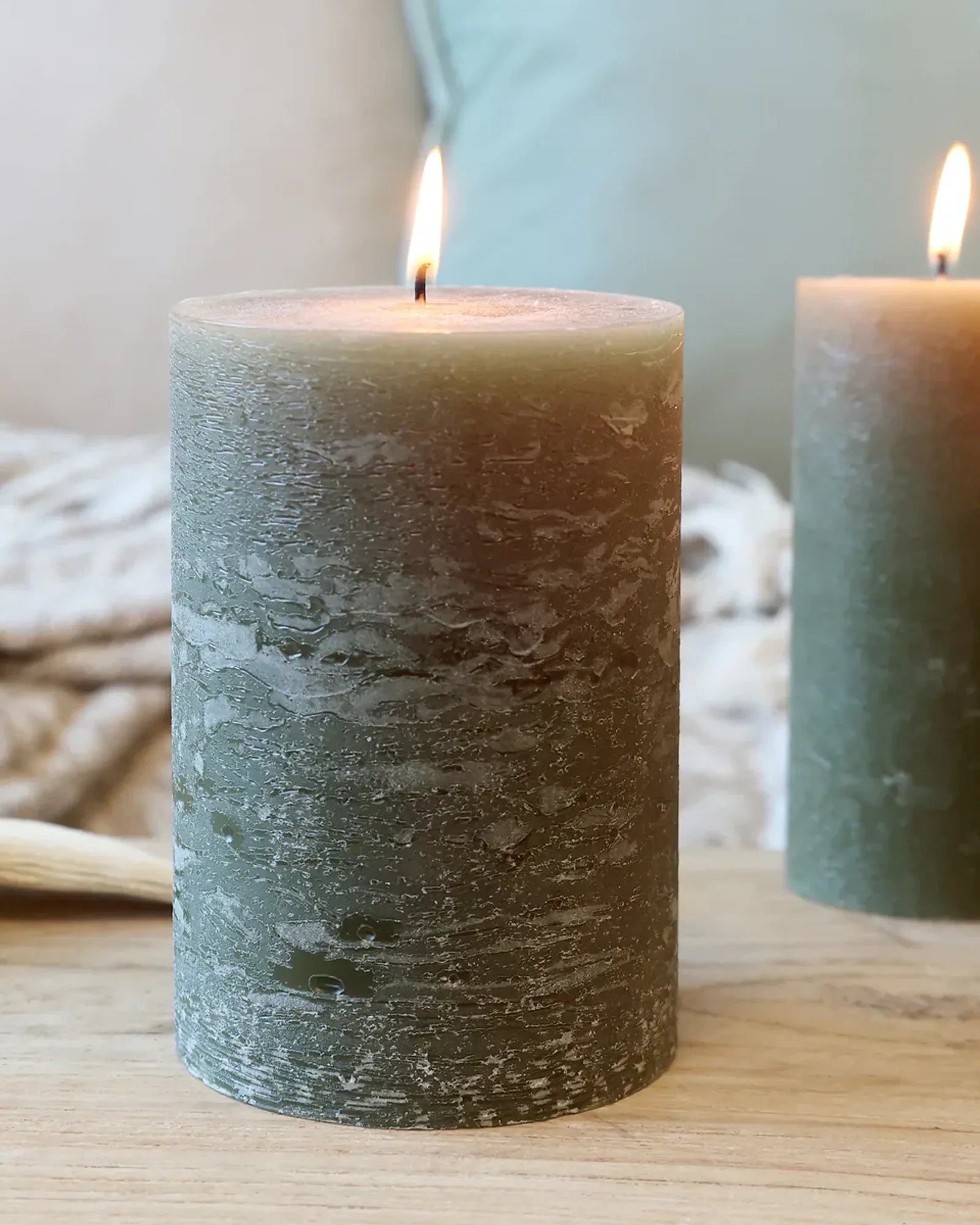 A wide rustic pillar candle in olive green, lit, with smaller candles of the same colour behind it.