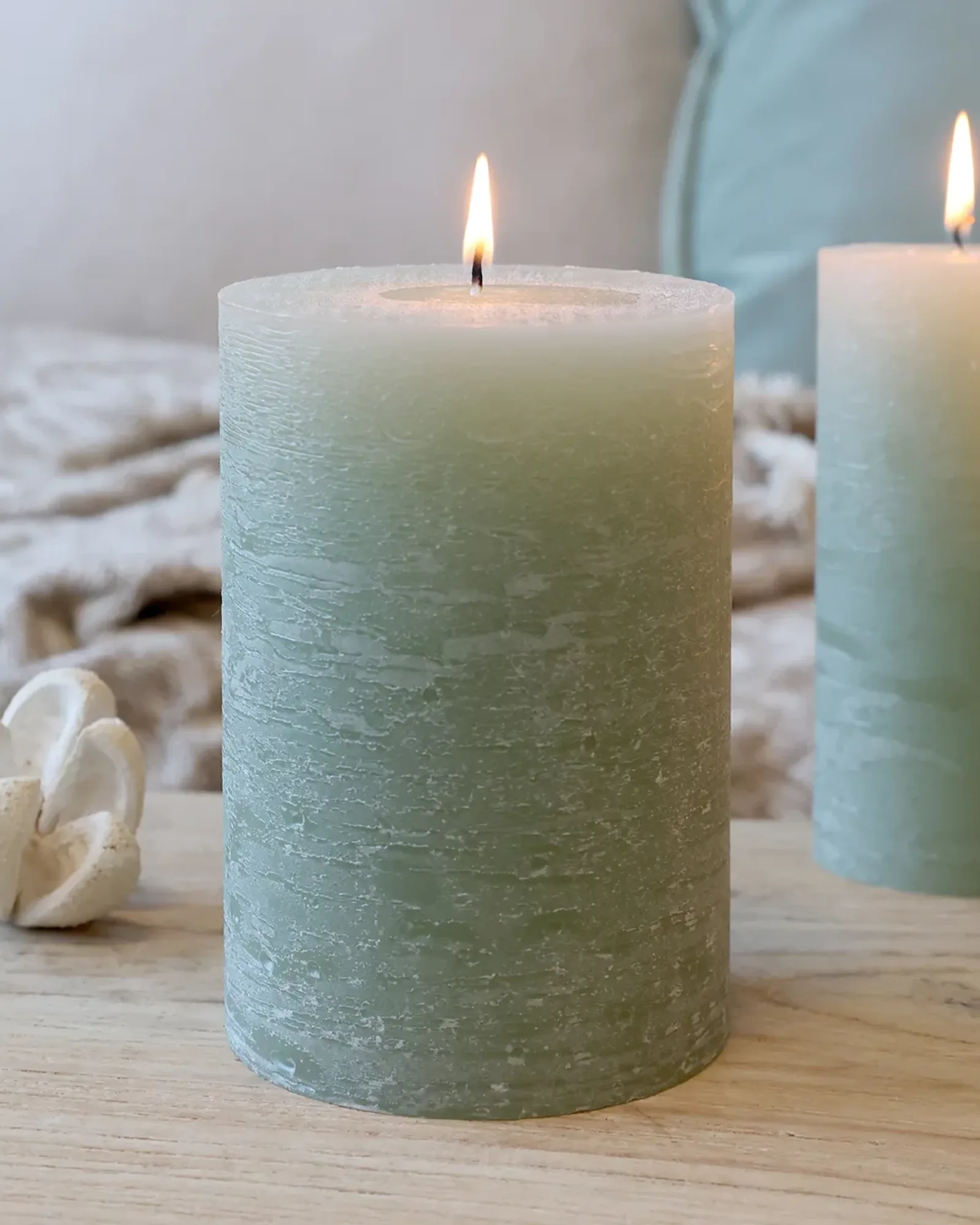 A wide rustic pillar candle in sage green, lit, with smaller candles of the same colour behind it.